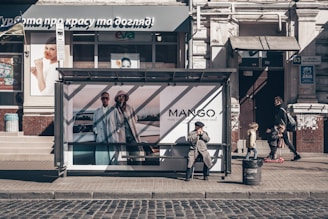 A city street featuring a bus stop with an advertisement for a fashion brand. An elderly man sits at the bus stop, wearing a coat and hat, with a thoughtful expression. Nearby, a man walks with two children, one on a scooter. The storefront in the background has various advertisements and a sign in a foreign language.