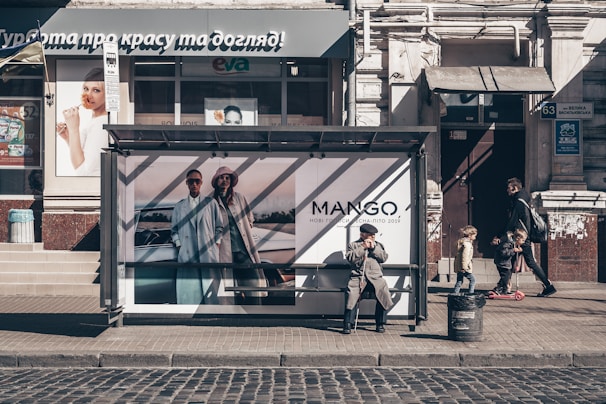 A city street featuring a bus stop with an advertisement for a fashion brand. An elderly man sits at the bus stop, wearing a coat and hat, with a thoughtful expression. Nearby, a man walks with two children, one on a scooter. The storefront in the background has various advertisements and a sign in a foreign language.
