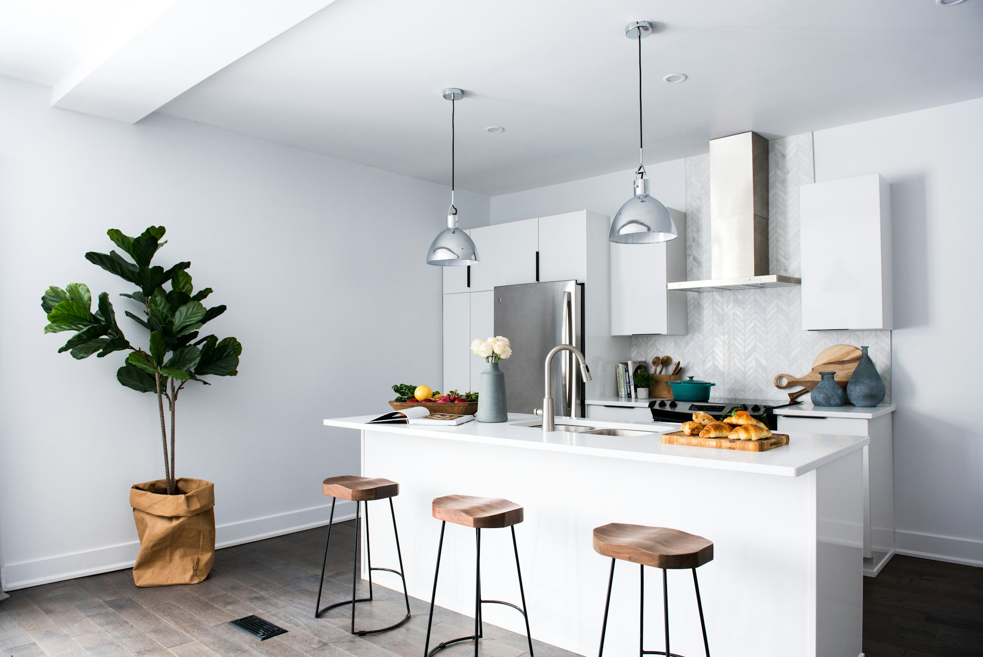 A bright, white, empty kitchen scene with wooden stools and a big green plant.
