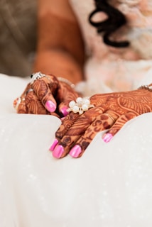 Intricately designed henna patterns adorn the hands, which are placed gently on a white, textured fabric. The nails are painted in a vibrant pink color, and a silver bracelet complements the overall look. A decorative ring featuring pearls adds elegance.