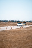 Two cars are driving on a paved track lined with orange traffic cones in a rural area. The background features trees and a clear sky, while the surrounding landscape consists of dry grass.