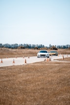 Two cars are driving on a paved track lined with orange traffic cones in a rural area. The background features trees and a clear sky, while the surrounding landscape consists of dry grass.