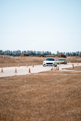Two cars are driving on a paved track lined with orange traffic cones in a rural area. The background features trees and a clear sky, while the surrounding landscape consists of dry grass.