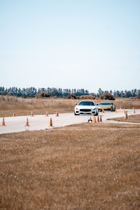 Two cars are driving on a paved track lined with orange traffic cones in a rural area. The background features trees and a clear sky, while the surrounding landscape consists of dry grass.