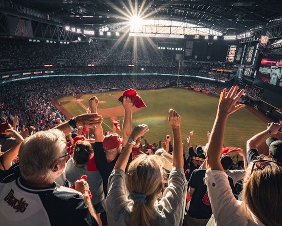 A lively crowd is cheering enthusiastically during a baseball game in a stadium. The sun is shining brightly through the partially open stadium roof, creating dramatic lighting. Fans are raising their hands with some holding red cloths, adding to the excitement of the scene.
