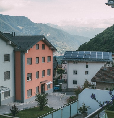A scenic view of a residential area with houses featuring colorful facades, surrounded by lush green hills. The house in the foreground has an orange color, while the one next to it has solar panels installed on the roof, reflecting a focus on renewable energy. In the background, a mountainous landscape stretches into the distance under a cloudy sky.