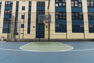 A basketball court is situated in front of a multi-story building with multiple windows. The court is enclosed by a tall chain-link fence, and a basketball hoop is visible. The building has a beige and blue color scheme with air conditioning units visible in some windows.