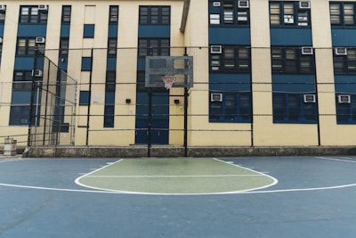 A basketball court is situated in front of a multi-story building with multiple windows. The court is enclosed by a tall chain-link fence, and a basketball hoop is visible. The building has a beige and blue color scheme with air conditioning units visible in some windows.