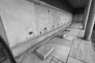 A row of stone basins with holes, likely for water, are built into a wall of light-colored stone. The foreground includes multiple stone seating areas, and the entire scene is framed by stone columns. The flooring and wall feature a consistent marble texture.