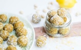 A collection of round, coconut-covered snacks displayed on a plate and in a glass jar. The treats are golden brown, suggesting ingredients like nuts or dates, and are sprinkled with shredded coconut. In the background, a lemon is partially visible, hinting at a fresh, citrusy flavor.
