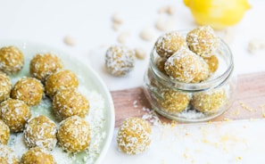 A collection of round, coconut-covered snacks displayed on a plate and in a glass jar. The treats are golden brown, suggesting ingredients like nuts or dates, and are sprinkled with shredded coconut. In the background, a lemon is partially visible, hinting at a fresh, citrusy flavor.
