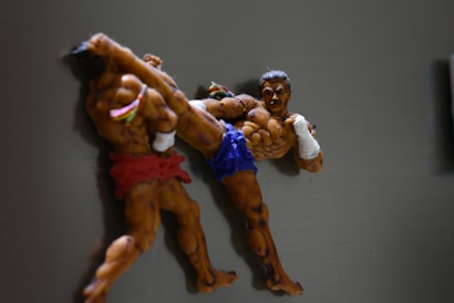 Two sculpted figures are captured in a dynamic martial arts pose. Both figures are muscular, with one wearing red shorts and the other in blue, executing a high kick. The details highlight their intense expression and body posture.
