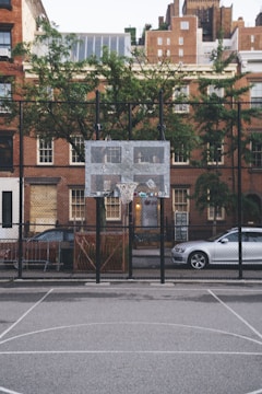 A city basketball court with a metal backboard and hoop is situated in front of an urban residential area with brick buildings. A tree is visible behind the fence that encloses the court. Parked cars line the street next to the buildings, and the scene conveys a quiet, urban environment.