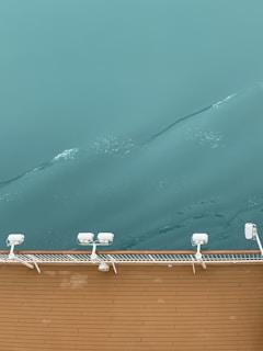 A section of a ship's deck with teak wood planks is bordered by a white railing equipped with several mounted lights. Beyond the railing, the calm, teal-colored water shows gentle ripples and light reflections, creating a serene maritime scene.