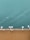 A section of a ship's deck with teak wood planks is bordered by a white railing equipped with several mounted lights. Beyond the railing, the calm, teal-colored water shows gentle ripples and light reflections, creating a serene maritime scene.