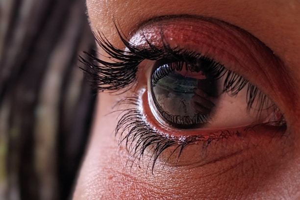 A close-up image of a human eye with detailed emphasis on the eyelashes and reflection visible on the eye surface. The reflection includes an indistinct view that could be a landscape or room.