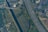 An aerial view of a sprawling urban area featuring a highway with multiple lanes, a river or canal running parallel, and roads lined with buildings and houses. The surrounding landscape includes patches of fields and trees, creating a contrast between the natural and built environment.
