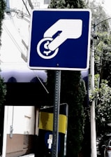 A blue and white sign depicts a hand inserting a coin into a slot or meter. The background includes a building, trees, and a utility pole with wires.