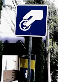 A blue and white sign depicts a hand inserting a coin into a slot or meter. The background includes a building, trees, and a utility pole with wires.