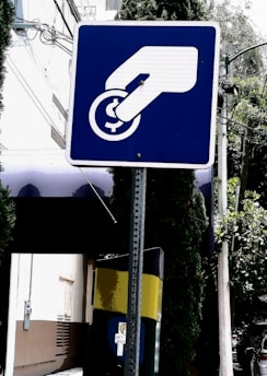 A blue and white sign depicts a hand inserting a coin into a slot or meter. The background includes a building, trees, and a utility pole with wires.