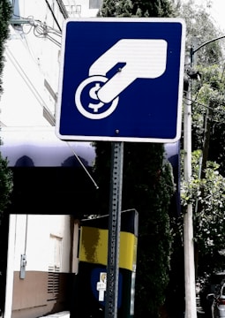 A blue and white sign depicts a hand inserting a coin into a slot or meter. The background includes a building, trees, and a utility pole with wires.