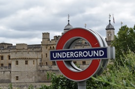 A prominent 'Underground' sign featuring a circular red frame with a blue bar displaying the word 'Underground'. In the background, an old stone fortress with multiple turrets and towers can be seen. The sky is overcast with gray clouds, and there is greenery in the foreground.