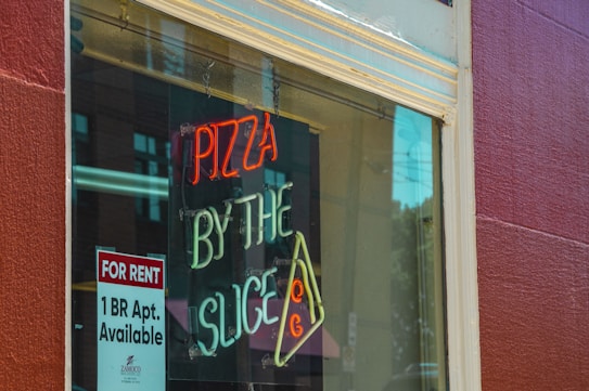A window display featuring a neon sign advertising 'Pizza by the Slice' with red and white lettering and a slice of pizza depicted in orange and yellow. To the side, a paper sign announces a 'For Rent' 1BR apartment available. Reflections in the glass reveal part of the street and buildings behind the viewer.