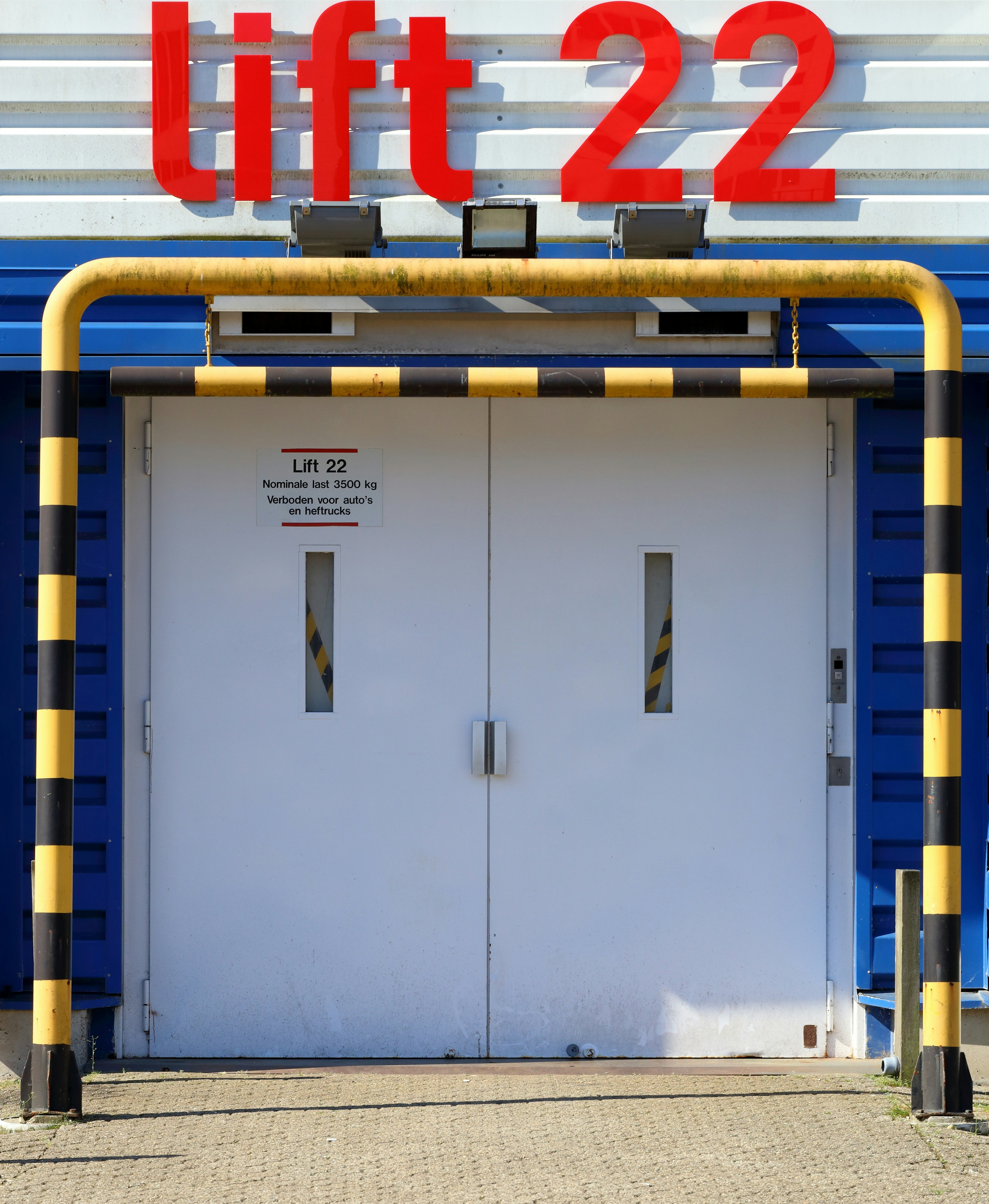 A large set of white industrial doors with handles, framed by sturdy blue metal. Above the doors, a bright red sign reads 'Lift 22' in bold letters. A yellow and black striped barrier runs horizontally above the doors, with matching striped poles on either side, adding a sense of caution. The ground is paved with concrete bricks.