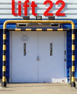 A large set of white industrial doors with handles, framed by sturdy blue metal. Above the doors, a bright red sign reads 'Lift 22' in bold letters. A yellow and black striped barrier runs horizontally above the doors, with matching striped poles on either side, adding a sense of caution. The ground is paved with concrete bricks.