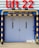 A large set of white industrial doors with handles, framed by sturdy blue metal. Above the doors, a bright red sign reads 'Lift 22' in bold letters. A yellow and black striped barrier runs horizontally above the doors, with matching striped poles on either side, adding a sense of caution. The ground is paved with concrete bricks.