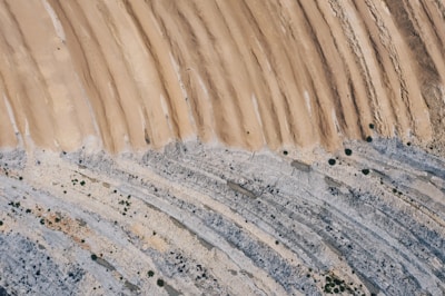 An aerial view of layered geological formations with a mix of sandy and rocky textures. The top portion features parallel ridges of a light brown sand, transitioning into patches of white and grey stone. Sparse vegetation is visible as small green dots scattered throughout the rocky areas.