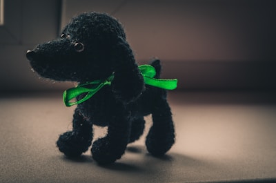 A small, black, knitted toy dog stands on a flat surface. It wears a bright green ribbon around its neck. The lighting creates a soft contrast, highlighting the texture of the knit material.