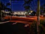 A street scene at night featuring a service garage with brightly lit interiors. Cars are parked outside, and the faint blur of car lights indicates motion. Foreground includes blurred palm leaves and a section of the road with arrow markings.