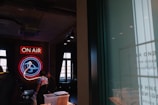 A neon sign with the words 'ON AIR' and a bird symbol illuminates a dimly lit room. The room has large windows and visible ceiling lighting. A person wearing a tie-dye shirt stands beneath the neon sign, surrounded by various objects and furniture.