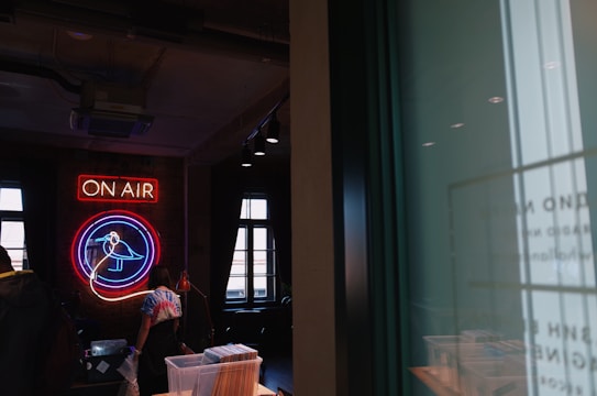 A neon sign with the words 'ON AIR' and a bird symbol illuminates a dimly lit room. The room has large windows and visible ceiling lighting. A person wearing a tie-dye shirt stands beneath the neon sign, surrounded by various objects and furniture.