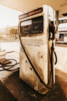 An old and weathered gas pump stands at a deserted gas station. The pump displays worn-out labels and dials, with a hose draped over the side. The background reveals a sunny day with soft, warm lighting, casting shadows on the pavement.