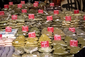 A market stall displays numerous bags of spices and herbs, each labeled with a price and name on red signs. The bags are neatly stacked, showcasing a variety of textures and colors. Some familiar spices such as mint, basil, and rosemary are visible.