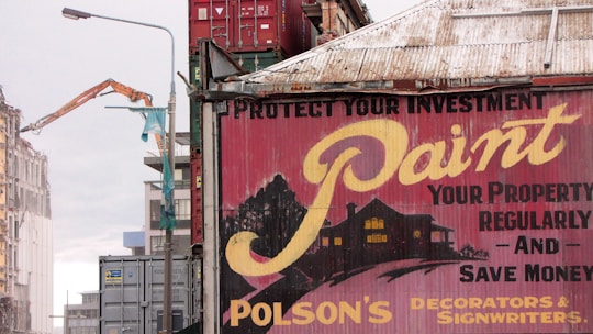 A large advertisement painted on the side of a building promotes painting your property regularly to save money, showing a rustic house silhouette. To the left, a construction site with a demolition crane indicates possible urban development. Shipping containers and a rooftop are visible, suggesting an industrial area.
