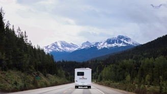 A scenic view of a mountainous landscape with snow-capped peaks and lush green forests lining the winding road. A large white RV is traveling on the road, heading towards the mountains. The sky is overcast with clouds, creating a serene and tranquil atmosphere.