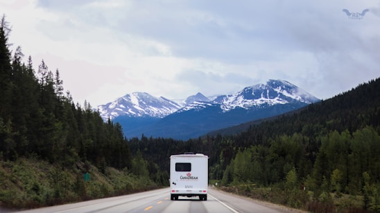 A scenic view of a mountainous landscape with snow-capped peaks and lush green forests lining the winding road. A large white RV is traveling on the road, heading towards the mountains. The sky is overcast with clouds, creating a serene and tranquil atmosphere.