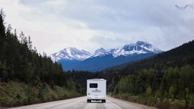 A scenic view of a mountainous landscape with snow-capped peaks and lush green forests lining the winding road. A large white RV is traveling on the road, heading towards the mountains. The sky is overcast with clouds, creating a serene and tranquil atmosphere.