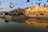 A historic fort is prominently featured in the image, sitting atop a hill and bathed in golden sunlight. Numerous birds are flying in the sky above the fort. Below, the fort and the birds are reflected in the calm waters of a large lake.