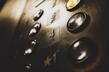 A close-up view of an elevator panel featuring several floor buttons with illuminated edges. The buttons have a metallic, reflective surface, and some display numbers while others have Braille markings. The golden hue of the lighting casts a warm glow over the panel.