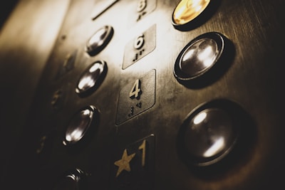 A close-up view of an elevator panel featuring several floor buttons with illuminated edges. The buttons have a metallic, reflective surface, and some display numbers while others have Braille markings. The golden hue of the lighting casts a warm glow over the panel.