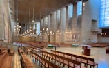 A spacious and modern cathedral interior with wooden pews arranged in rows. High walls feature tall vertical windows letting in natural light, and the ceiling is adorned with multiple hanging light fixtures. The altar area is open and minimally decorated, with flower arrangements and a lectern visible. Artwork depicting religious figures is displayed on the walls.