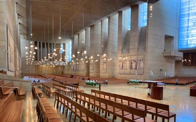 A spacious and modern cathedral interior with wooden pews arranged in rows. High walls feature tall vertical windows letting in natural light, and the ceiling is adorned with multiple hanging light fixtures. The altar area is open and minimally decorated, with flower arrangements and a lectern visible. Artwork depicting religious figures is displayed on the walls.