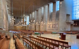 A spacious and modern cathedral interior with wooden pews arranged in rows. High walls feature tall vertical windows letting in natural light, and the ceiling is adorned with multiple hanging light fixtures. The altar area is open and minimally decorated, with flower arrangements and a lectern visible. Artwork depicting religious figures is displayed on the walls.
