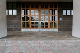 A building entrance with large glass-pane windows and two sets of double doors made of wood. The surrounding walls are made of dark, square tiles, and the floor consists of light-colored brick pavers arranged in a pattern.