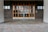 A building entrance with large glass-pane windows and two sets of double doors made of wood. The surrounding walls are made of dark, square tiles, and the floor consists of light-colored brick pavers arranged in a pattern.