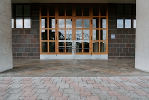 A building entrance with large glass-pane windows and two sets of double doors made of wood. The surrounding walls are made of dark, square tiles, and the floor consists of light-colored brick pavers arranged in a pattern.